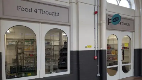 The front of the shop has two big cylinder shaped windows. The walls are brick and are painted half white and half black. You can see trolleys inside with food items and others on it. 