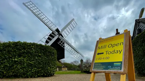 Ant Saddington/BBC A black-bodied wooden mill in 2023, with its white sails open in an X shape. In front of it is a yellow sign saying 'mill open today'