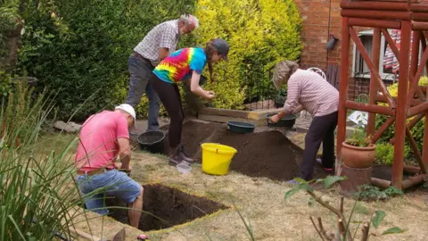 A view of four people digging a hole in a garden. One man in a pink t-shirt is inside a square hole, while three other people look through a pile of dirt.