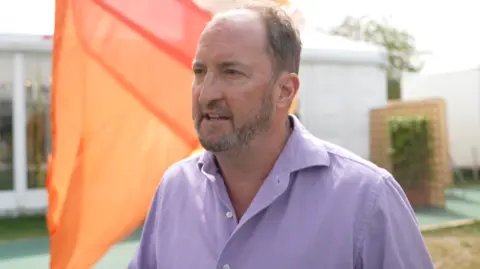 Guto Harri is facing sideways to the camera, wearing a lavender shirt, stood on the Hay Festival grounds.