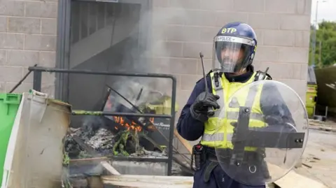 Getty Images A dwindling fire of wood and melted plastic in the doorway of a hotel. A british transport police officer with riot gear stands to the foreground.