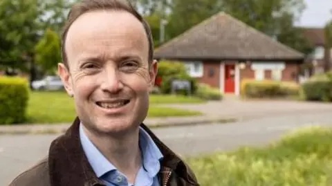 Richard Rout standing on a verge at a T-junction. He is smiling at the camera and wearing a brown wax jacket with a blue shirt underneath. In the background you can see the grass of the verge, some housing and the road markings of the junction. 