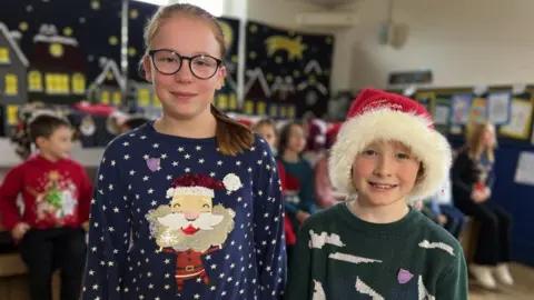 BBC Sophia and Seth from Castle Primary School in Stoke-sub-Hamdon in Somerset. Sophia is wearing a navy blue Christmas jumper with Santa on the front. Seth is stood next to her, wearing a red and white fluffy Santa hat and a green jumper. They are both looking at the camera and smiling. A Christmas display and other children can be seen behind them.