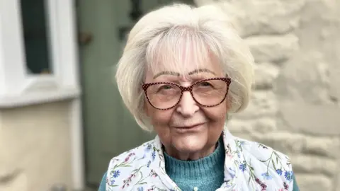 A white lady with white hair and red polka dot framed glasses wearing a flowery Gilet