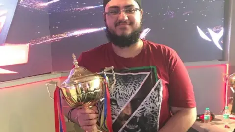 Ishpreet Singh Chadha A young Asian man with a bushy beard, glasses holds a large golden trophy. He's wearing a patka - a religious head covering worn by Sikh men. It sits tight on the head and has a topknot that sits on the crown of the skull. The trophy has elaborate handles on either side and blue and white ribbons dangling off them. The man's smiling, and wears a red t-shirt with a white tiger face design on it. The picture is cut in half diagonally, and underneath the face of a male is visible.