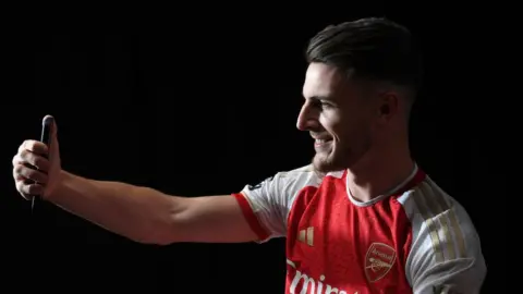 Getty Images Footballer Declan Rice sitting side on and holding a mobile phone in his right hand with his right arm outstretched as if he is taken a selfie picture or video of himself. He is smiling and has his black hair slicked back over his head. He is wearing the Arsenal home kit which is mainly red with white sleeves. The background behind him is totally black.