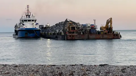 East Devon District Council A blue and white ship is on the left with piles of rocks on a barge next to it. There are also two pieces of plant machinery on the front of the barge. You can see the sea and the shore against a clear crepuscular sky.