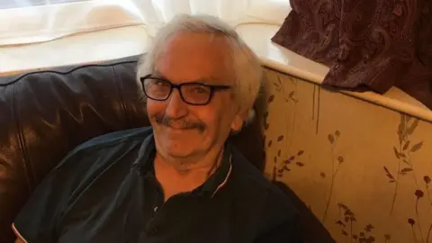 Terry Hall sits on a brown leather sofa. With white hair and a dark grey moustache and wearing a navy polo shirt.