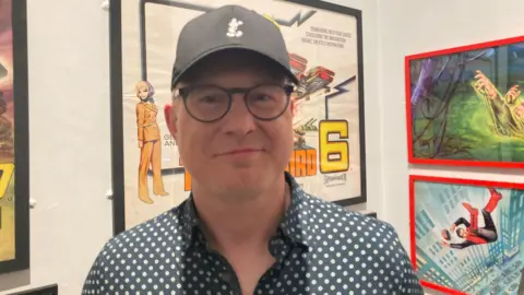 A man wearing glasses and a black cap standing in front of framed Thunderbirds artwork in a museum.