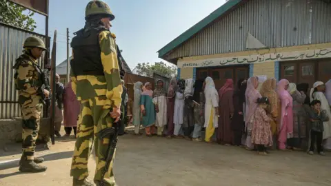 Getty Images Indian security personnel stand guard as voters queue to cast their votes at a polling station during the third and final phase of voting for local elections on October 1, 2024 in Bandipora.