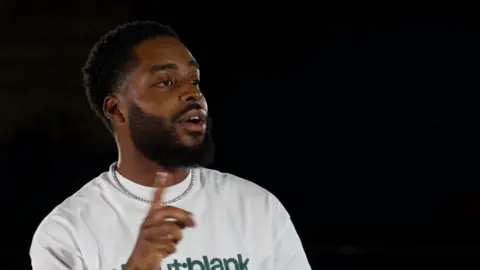 Only Zizou is mid conversation talking to a person out of frame on his right. He is wearing a silver chain around his neck and white crew neck T-shirt with the words 'About:Blank' written in green print on the centre. He has a black beard, black hair and a slit in his right eyebrow. He is stood in front of a black background.