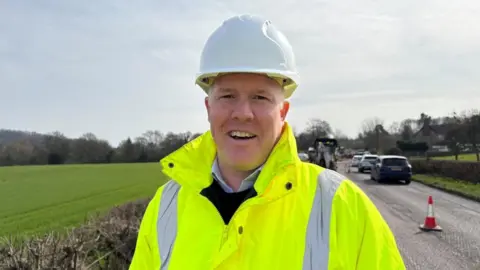 A man in a bright fluorescent yellow coat and a white hard hat is standing on a road and smiling at the camera. Behind him, there are cones down the centre of the road and cars going one way. On the opposite side of the road, in the distance, there is a large tractor. The road is bordered by hedgerows and green fields.