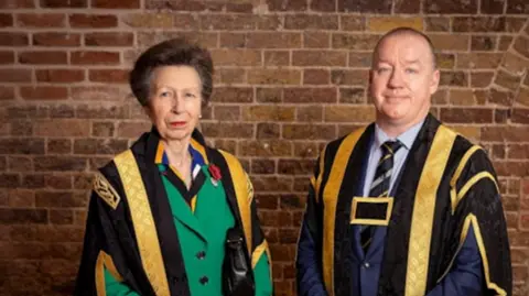 Princess Anne in black and gold robes and a green jacket stood next to a man wearing a black and gold robes and a blue suit, standing in front of a brick wall