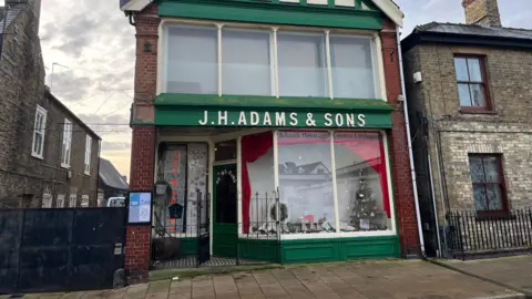 An old-fashioned shop from the Victorian era. It has dark green panelling and two large glass shop fronts, one on the second floor. It bears the name "JH Adams & Sons" in white capital letters.