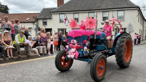 A woman drives a small tractor through a village. She is dressed entirely in pink and has decorated her tractor with pink decorations, toys and more. People sit on the side of the road clapping and cheering.