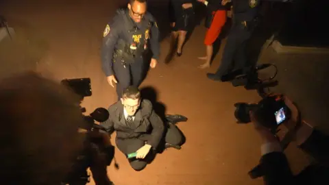 A man in a dark coloured suit sits on the floor among a group of people. There is a bright light focused on his face and a uniformed police officer stands behind him. 