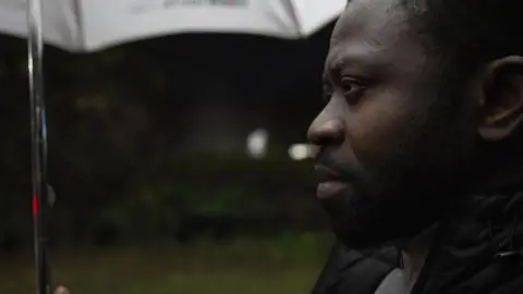 A close up of Mr Morakinyo, who is looking serious, wearing a black coat and holding an umbrella.
