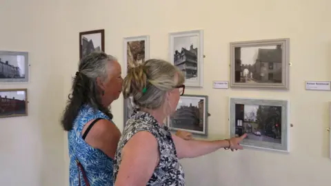 Two women are looking at photos, which are mounted on a wall in an exhibition, with archive photographs placed next to modern day re-creations of the same images.