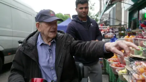 Mr Chafer points at some fruit and vegetables outside a shop, with an assistant standing behind him looking on
