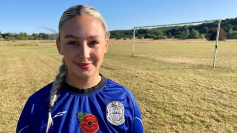 A woman in blue football shirt stands on a grass roots football pitch. She has blonde hair tied in a braid. Her shirt displays the club badge of Stoke Town Football Club, a poppy and a clothing logo. Rusty goal posts and trees can be seen in the distance.