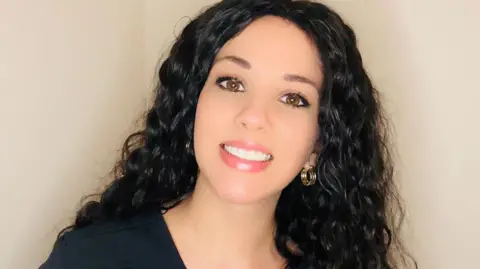 Anastasia, with long, curly black hair, is smiling at the camera and standing against a blank wall