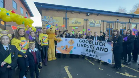 Olivia Courtney-Ashton/BBC A large group of schoolchildren, pictured with Pudsey the Bear, hold aloft a sign which reads: "Good luck Lindsey and Amy #teamairedale