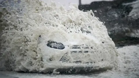 Getty Images A car drives through sea foam whipped up by the wind of Storm Ophelia at Trearddur Bay, Holyhead