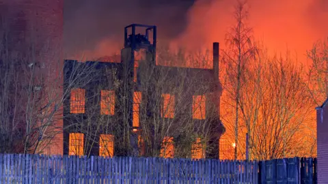 BBC A large blaze at night in a mill building. The sky is lit up orange. Wooden fencing is in front of the building.