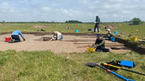 An archaeological dig is in progress with a wide, square shallow trench in which four people are digging while sitting or kneeling. They each have a brightly coloured bucket. A man is standing taking notes on the far side of the trench and in the foreground of the shot are some spades.