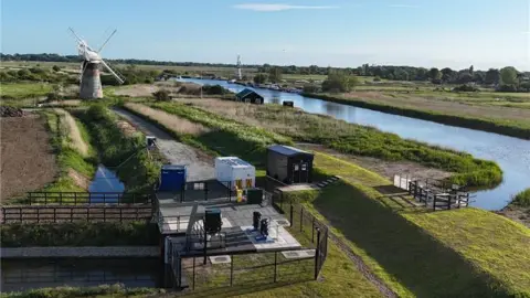 Water Management Alliance A drone image of a pumping station in the Broads, showing a modern electric station in the foreground, in front of an old windpump, next to the River Thurne
