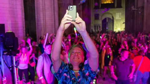 Peterborough Cathedral Paul Stainton smiles as he holds a smartphone above his head to take a picture of himself with hundreds of dancers behind him. He is smiling and wearing a short-sleeved, Hawaiian-style shirt. Cathedral walls can be seen.