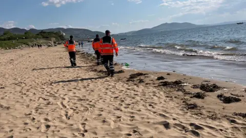 BBC Three people in high viz walking along a shoreline.