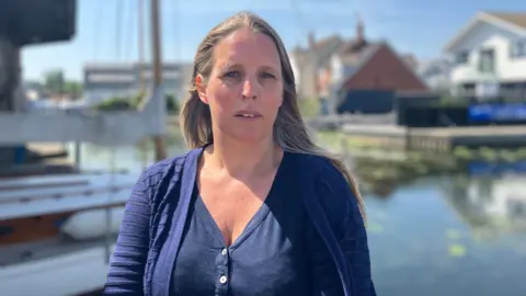 Owen Sennitt/BBC A woman with long blond hair looks towards the camera in front of a boat moored up at a boatyard in the Norfolk Broads on a sunny day. 