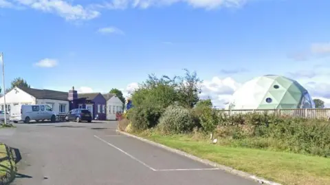 Google A road leads to white and purple buildings of The 68 cafe, which resemble bungalows. There is a sage green dome behind a fence on the right side of the road.