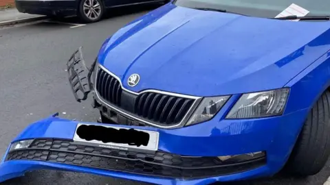 East of High St Residents A blue car is parked on a street with most of the bumper hanging off and damage to the side of the vehicle
