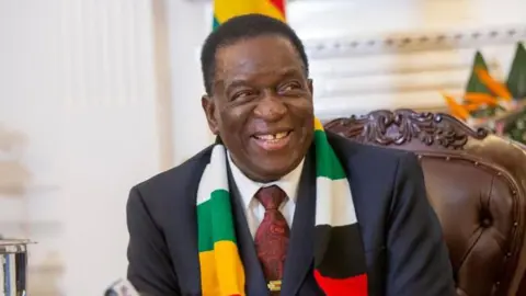 EPA-EFE President Emmerson Mnangagwa sits smiling wearing a dark suit, red patterned tie and a scarf in green, white, red and black colours of the Zimbabwean flag.  
