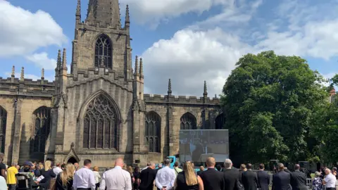 A crowd of people, many dressed in black, are stood outside Sheffield Cathedral watching a large video screen showing a funeral