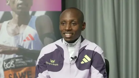 Sabastian Sawe sitting during an interview, wearing a lavender-coloured training top with a curtain behind him