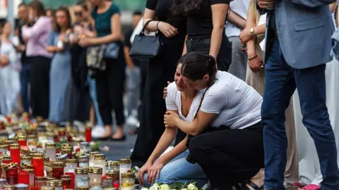 Matej Povse/Getty Images Two women in white T-shirts comfort each other amid a group of people and candles on the ground