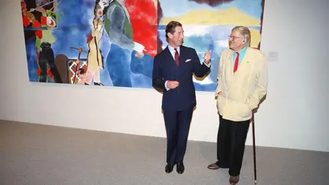 AFP via Getty Images David Hockney next to King Charles in 1994. Charles wears a dark suit, while Hockney is dressed in a white blazer, red tie and blue shirt, with black trousers. Behind them is a colourful piece of art