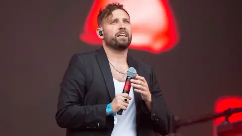 Getty Images You Me At Six's Josh Franceschi performing at Reading festival - he is wearing a black blazer, a white t-shirt and is holding a microphone