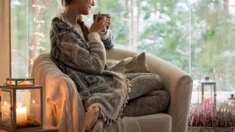 Getty Images Stock image of woman wrapped up in knitwear with fairy lights and a hot drink