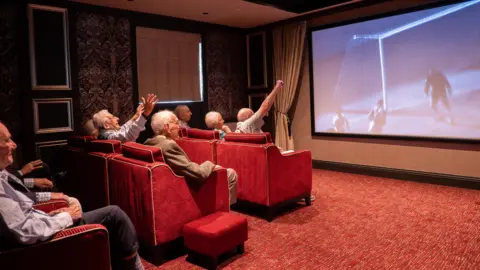 Hendon Hall Care Home Residents and guests cheer on England again whilst watching the 1966 World Cup final. They are sitting in comfy red chairs and looking at a big screen