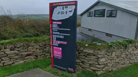 Google Maps The sign at the entrance of Helston Business Park which has a map of the businesses and a key to the businesses' names. There is a corrugated large building in the background and in the foreground is a stone wall and grass.