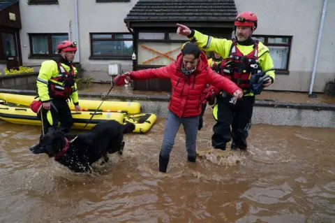 PA Media Members of the emergency services help a woman in Brechin, Scotland, as Storm Babet batters the country.