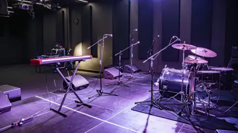 Access Creative College A stage with an electric keyboard, microphones, drum kit and speakers with professional lighting.