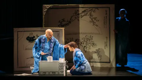 Mihaela Bodlovic A man and a woman wearing blue robes kneel on the stage. There is a small kitchen unit on the stage and two boards in the background with black ink drawings, inspired by Japan.