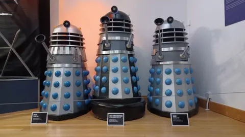 A group of three prop Daleks, conical robotic alien creatures, from the BBC science-fiction series Doctor Who, on display as museum exhibits against a white background. The two on either side are silver with blue trim, while the one in the centre stands slightly taller on a large black fender, with black stripes and a black dome section at the top in addition to the silver and blue of the others.