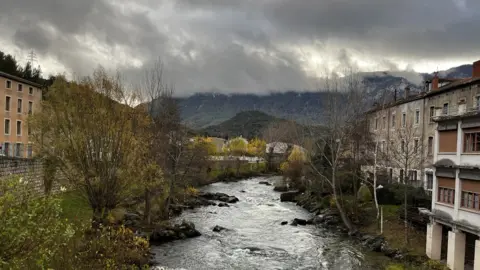 BBC A view of the French town of Quillan. A river is seen with houses on each bank. In the background are some cloud-covered mountains.