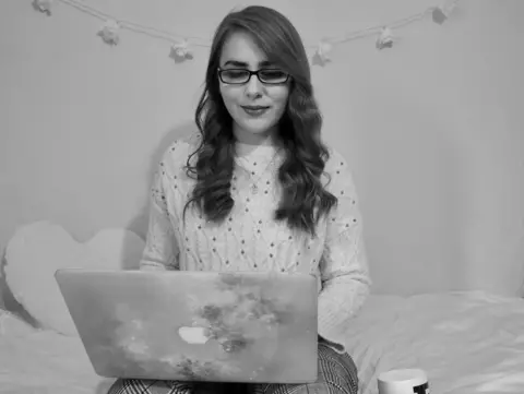 Williams family Elin sitting on a bed with a love heart cushion behind her, working on a laptop